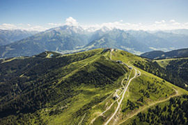 Panoramablick auf den grünen Gipfel der Schmittenhöhe sowie eine schmale Straße, die sich durch die Berglandschaft zieht, mit Bergen im Hintergrund. © (c) Zell am See-Kaprun Tourismus Panoramablick auf den grünen Gipfel der Schmittenhöhe sowie eine schmale Straße, die sich durch die Berglandschaft zieht, mit Bergen im Hintergrund.
