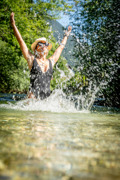 Eine Frau in schwarzem Badeanzug und Strohhut spielt fr&ouml;hlich im klaren Wasser eines Flusses. Mit erhobenen Armen spritzt sie Wasser in die Luft, Sonnenlicht funkelt auf den Tropfen. Im Hintergrund sind gr&uuml;ne B&auml;ume und Berge unter blauem Himmel zu sehen.