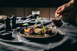 Hand gie&szlig;t dunkle Sauce aus einem kleinen Glas-K&auml;nnchen &uuml;ber einen kunstvoll angerichteten Teller mit Lammkarree, Gem&uuml;se und Kartoffelp&uuml;ree auf einem gedeckten Tisch.