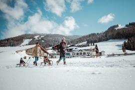 Familie zieht zwei Kinder auf Schlitten durch die verschneite Winterlandschaft, im Hintergrund Hotelgeb&auml;ude und Skipisten unter blauem Himmel.