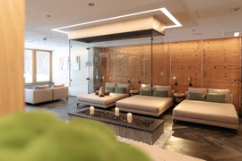 Heller Ruheraum im Spa mit gro&szlig;en Liegen, Kissen und Kerzen in einer Feuerstelle, umgeben von Holzverkleidung und Glasw&auml;nden.