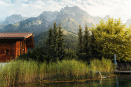 Naturbadeteich im Sommer, umgeben von gr&uuml;nen Str&auml;uchern und B&auml;umen, daneben das Badhaus, im Hintergrund Bergkulisse.