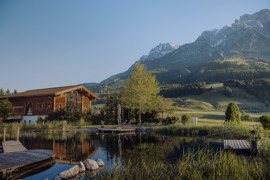 Naturbadeteich im Sommer, umgeben von gr&uuml;nen Str&auml;uchern und B&auml;umen, im Hintergrund das Badhaus, gr&uuml;ne Wiesen und Berge.