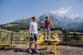 Mann hilft einem kleinen M&auml;dchen beim Balancieren auf dem Abenteuerspielplatz im Sommer, im Hintergrund gr&uuml;ne Wiesen und Bergkulisse.