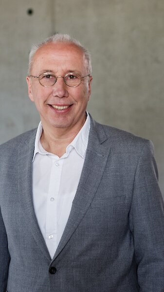 Portraitbild Dr. Thomas Belazzi. Mann zwischen 50 und 60 Jahren mit schütterem Haar, Brille, grauen Anzug und weißem Hemd, er lächelt.