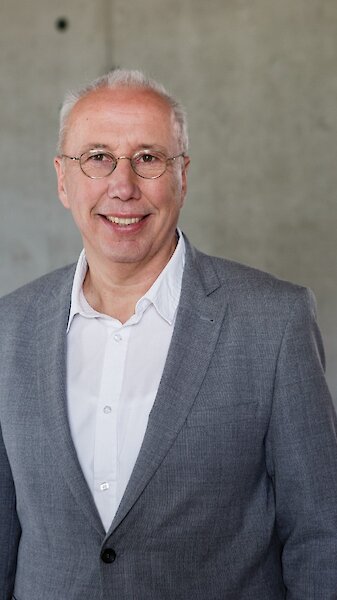 Portrait picture of Dr Thomas Belazzi. Man between 50 and 60 years old with thinning hair, glasses, grey suit and white shirt, smiling. Copyright by Pletterbauer.